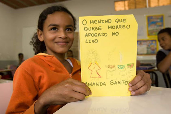 A smiling student holds up a yellow book in a classroom.