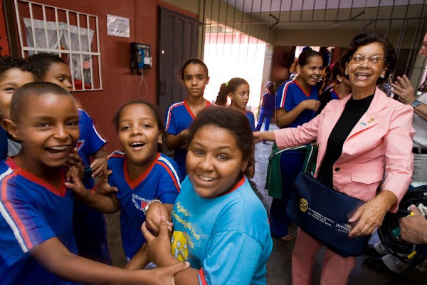 Smiling schoolchildren gather beside an adult visitor outside a school.
