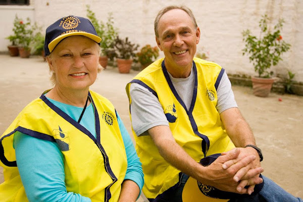 Two smiling volunteers wearing yellow Rotary vests sit together outdoors.